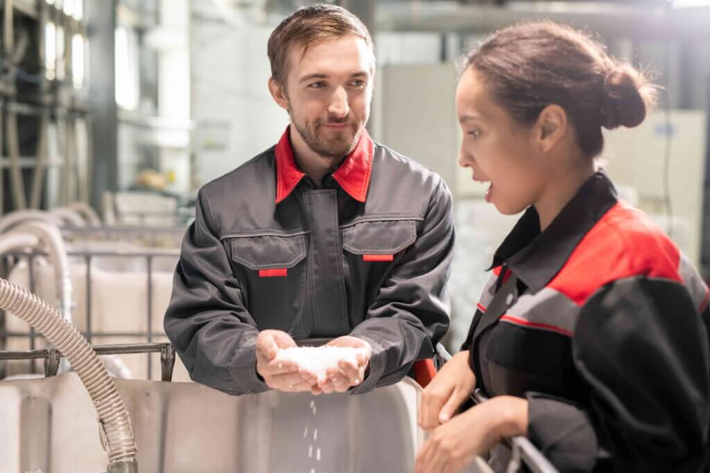 Two young quality control managers discussing characteristics of white plastic pellets while one of them holding pile over large container.