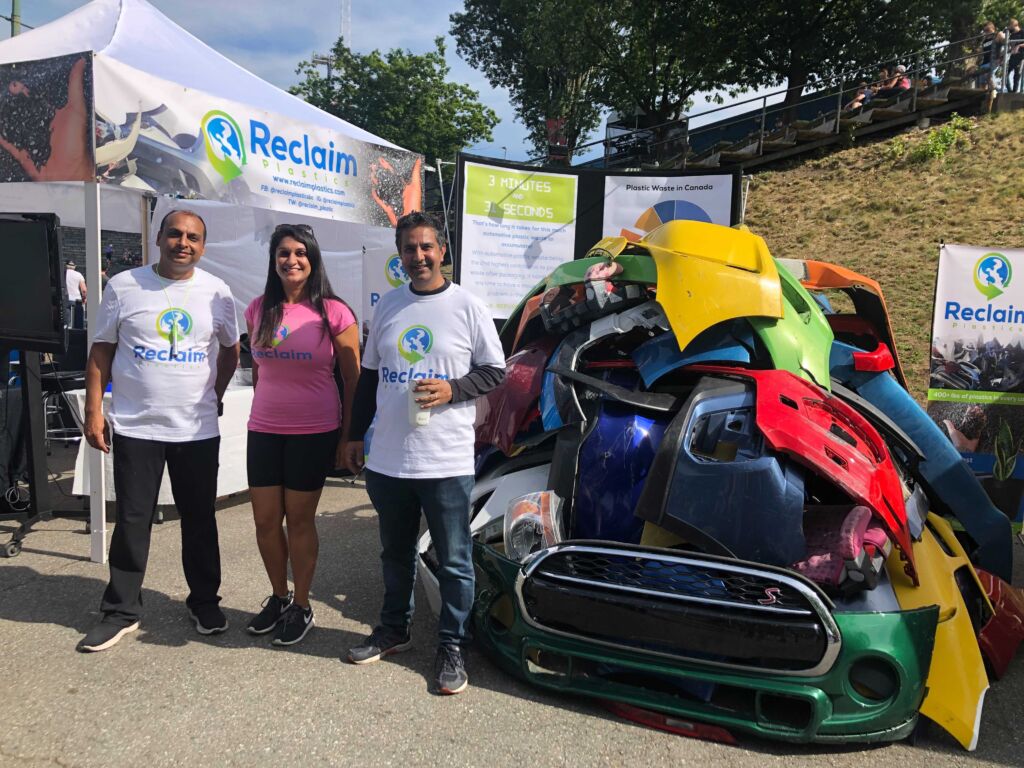 The photo shows three people standing between a pile of damaged car bumpers and the Reclaim Plastics booth outside at the PNE in Vancouver, BC.