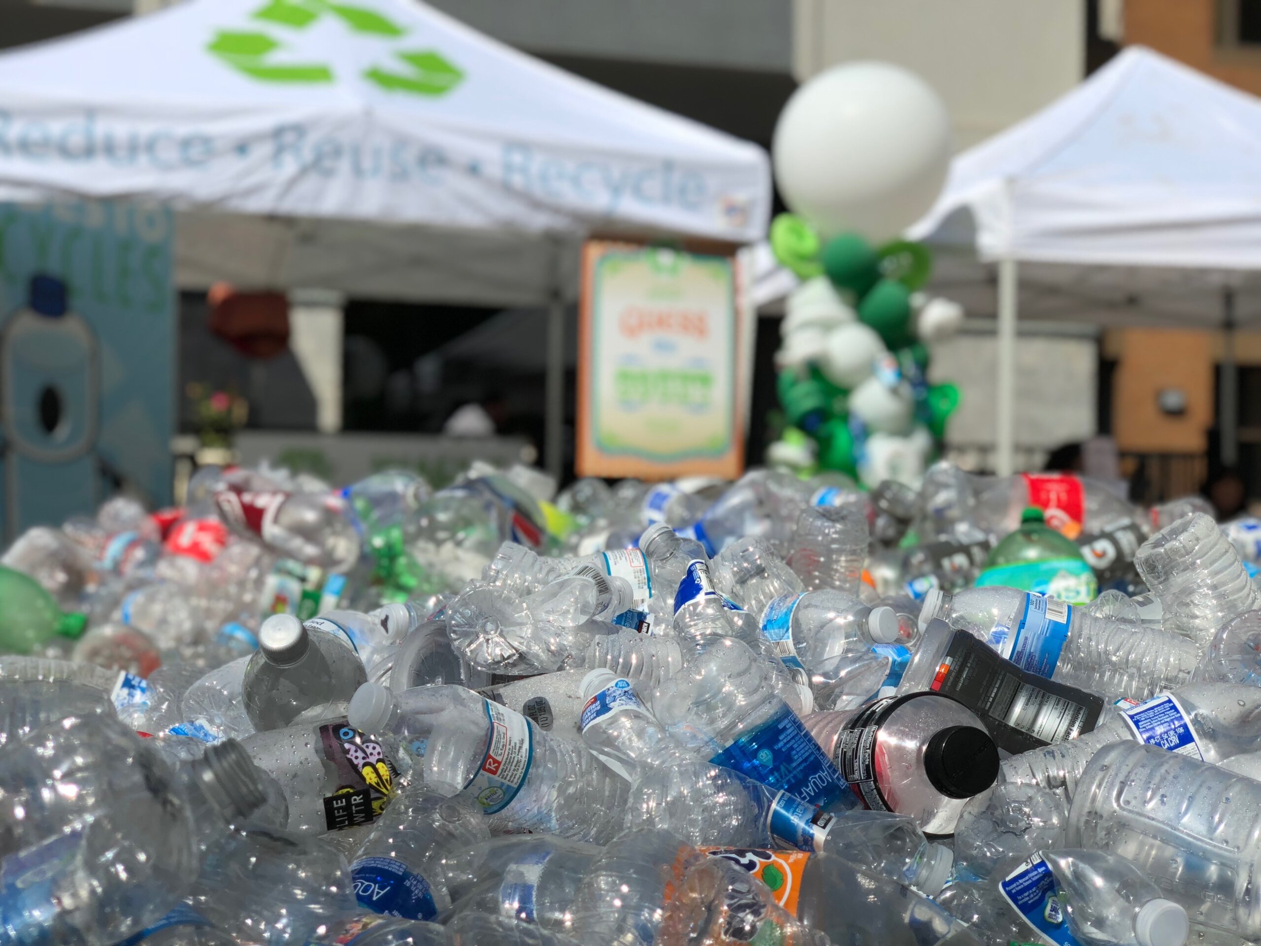 A pile of plastic bottles in the foreground with a tent in the background that reads "reduce, reuse, recycle" on the canopy.