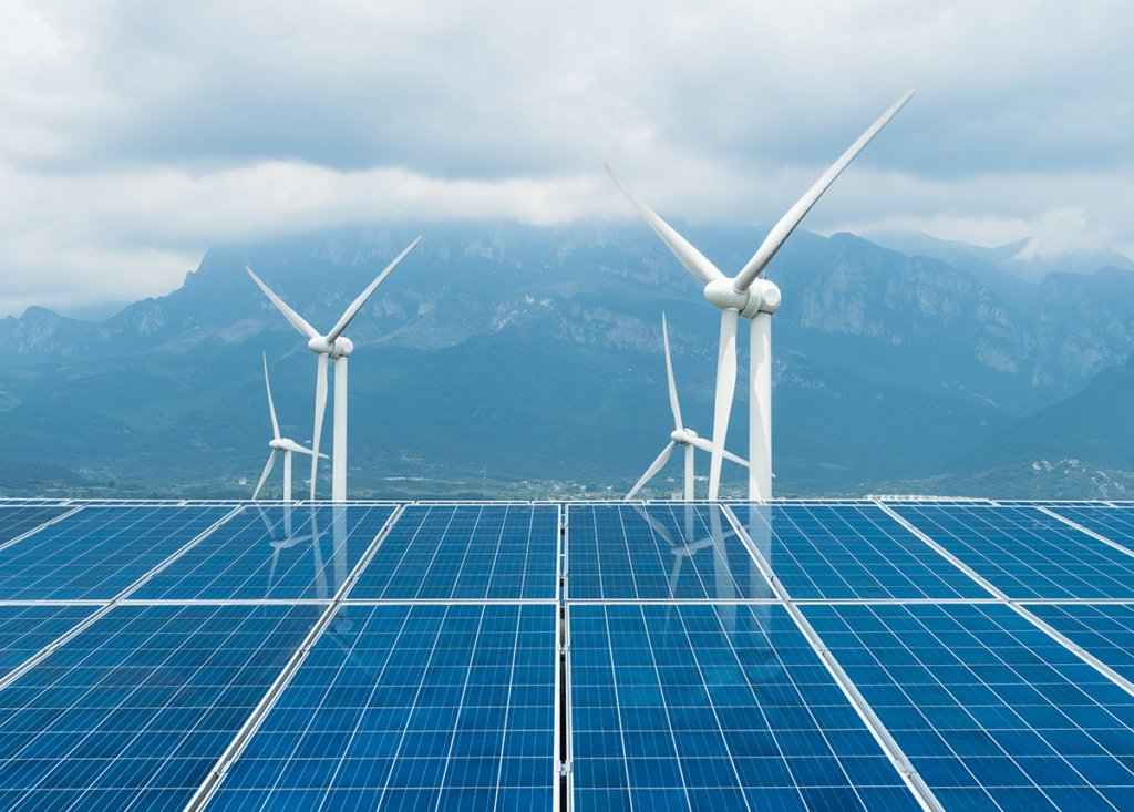 A photo of a cloudy mountain landscape with four wind turbines in the background and solar panels in the foreground.