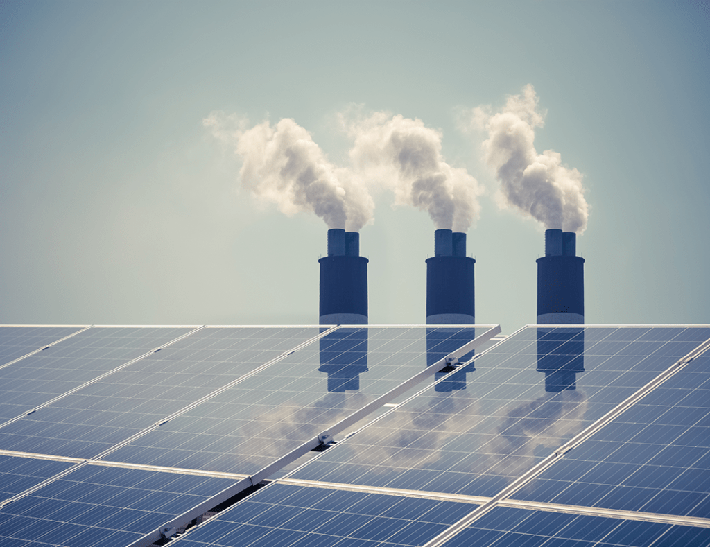 A photo of three chimneys blowing smoke against a blue sky, with solar panels in the foreground to symbolize a clean energy transition.